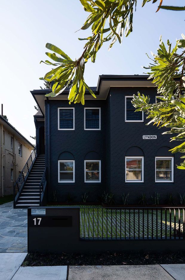 Street-facing view of a dark charcoal two-storey residential building with a sharply articulated low-pitched roof, textured exterior walls, white-trimmed windows, and an external stair along the side. A black metal slat fence, paved front path, letterbox, and address number 17 define the foreground, while leafy branches partially frame the facade.