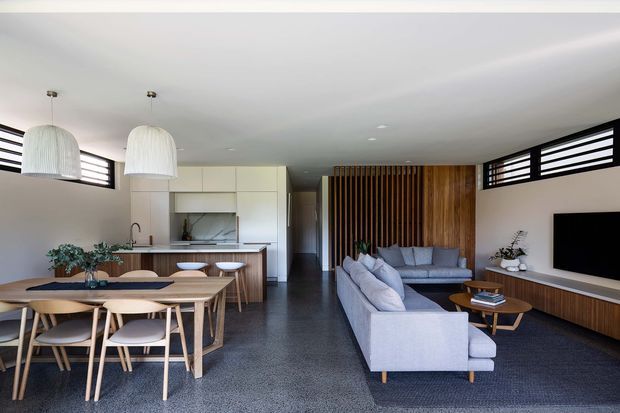 Open-plan contemporary living, dining and kitchen space with a neutral palette and strong timber detailing. The room features a large light-grey sectional sofa, timber dining table and chairs, white kitchen cabinetry with island seating, a dark textured floor, horizontal clerestory-style windows, pendant lights, and a timber slatted feature wall behind the living area.
