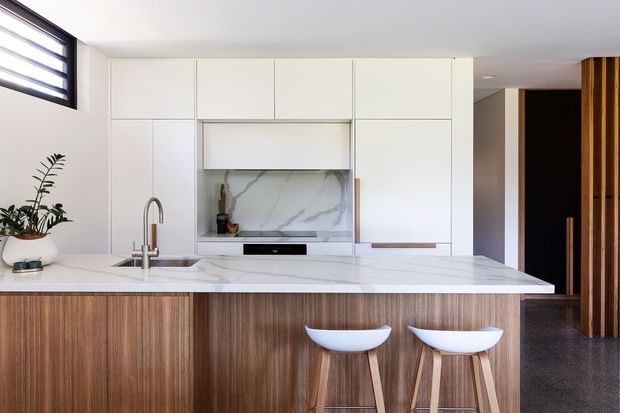 Contemporary open-plan kitchen dominated by a large island with vertically grained timber cabinetry, a white engineered-stone benchtop, integrated white wall cabinetry, and a veined stone splashback. Two white sculptural bar stools sit at the island. A dark hallway and vertical timber screening are visible beyond the kitchen, with a small high horizontal window providing natural light.