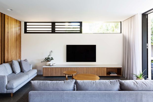 A bright contemporary living room with a large grey sectional sofa in the foreground, a matching side sofa, and a low timber entertainment console beneath a wall-mounted television. Vertical timber wall panelling forms a feature wall on the left, while high horizontal windows, full-height glazing, and sheer curtains bring in abundant daylight. A pair of simple timber coffee tables and restrained greenery complete the calm, refined interior.