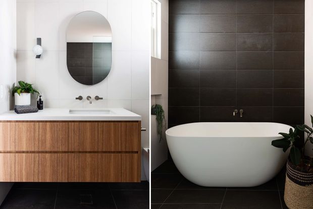 A refined contemporary bathroom featuring a wall-mounted timber vanity with a white benchtop and integrated basin, paired with brushed metal tapware and an arched black-framed mirror. A freestanding white oval bathtub sits against a dark tiled feature wall, with matching dark floor tiles, white wall panels, greenery, and understated decorative accessories.
