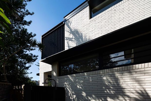 Low-angle exterior view of a contemporary multi-level building with crisp white brick cladding, strong black projecting roof and balcony elements, dark vertical screening, and broad dark-framed horizontal glazing. Tree shadows fall across the facade, with mature landscaping visible at the side.