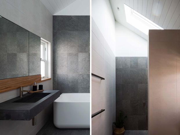 A contemporary bathroom shown in a split view. The space features large dark grey stone-look wall and floor tiles, a black vanity with integrated basin and wall-mounted tapware, timber shelving, a freestanding white bathtub, and a large mirror. The second view shows a high white paneled ceiling with a skylight, tiled shower zone, and a warm metallic-toned privacy panel. The palette is monochromatic with timber accents and abundant natural light.