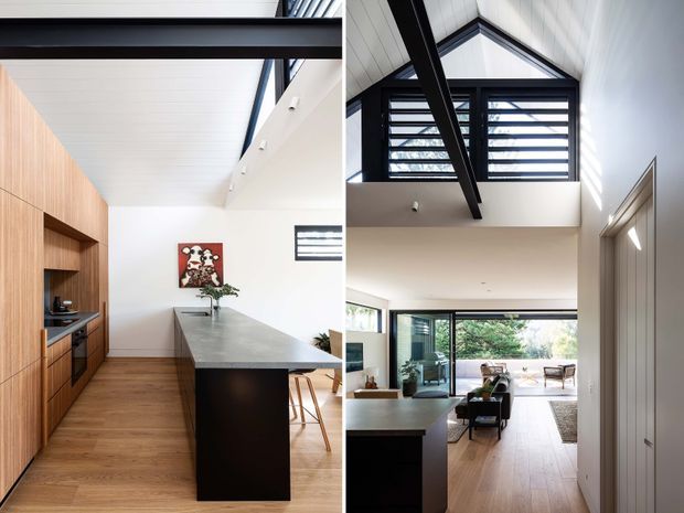 A bright, contemporary open-plan kitchen and living area with a dramatic double-height void. The kitchen features warm timber cabinetry, a dark island base with a grey stone benchtop, timber flooring, and white wall and ceiling finishes. Black exposed structural beams and framing contrast with extensive glazing and high-level louvred windows. The adjacent living space opens to a leafy outdoor area.