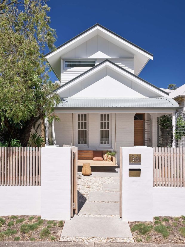 Front exterior view of a bright white two-storey gabled house with layered pitched roofs, horizontal weatherboard cladding, a corrugated metal verandah roof, and a small fenced front garden. The entry is approached through a pedestrian gate and gravel-and-concrete path, with timber doors and glazed French-style front doors visible beneath the verandah.