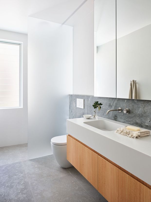 Bright contemporary bathroom with a floating timber vanity, integrated white benchtop and basin, large mirrored wall cabinets, grey stone-look splashback and floor tiles, wall-mounted tapware, and a concealed toilet. A frosted window provides soft natural light.