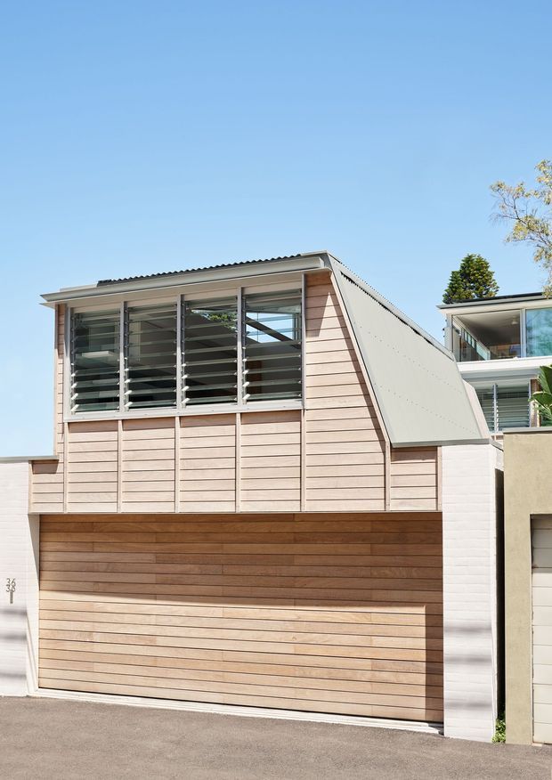 Street-facing view of a compact contemporary residential garage and upper-level living space. The facade combines horizontal timber-look cladding and a timber garage door with white masonry side walls, a steep skillion-like roof form, and broad louvred windows beneath a projecting roof edge. A neighbouring modern residence is partially visible at right.