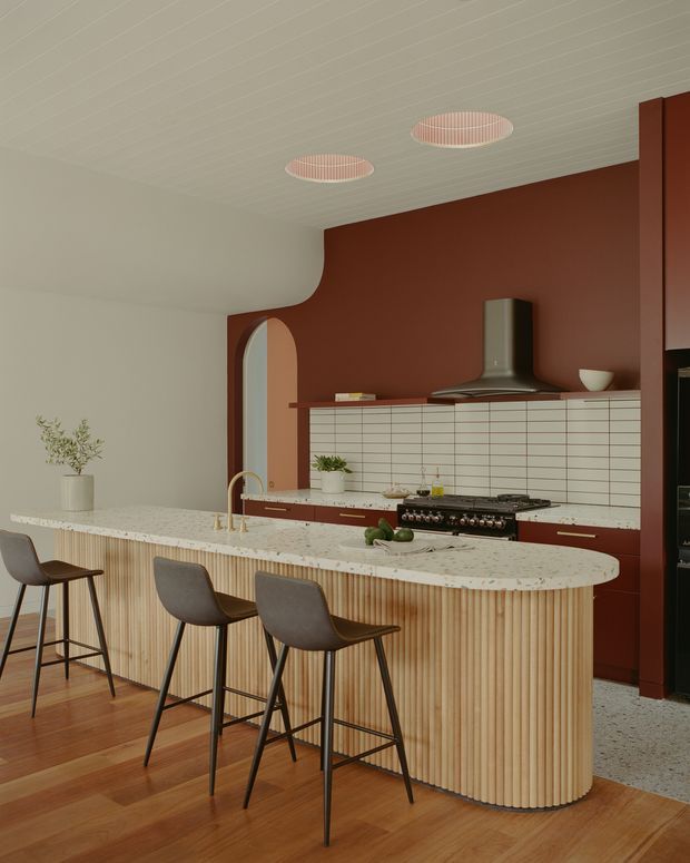 A warm, refined kitchen with a large curved island featuring vertically fluted timber cabinetry and a pale stone benchtop. Three dark upholstered bar stools sit along the island. The rear wall is painted deep terracotta-red and fitted with a white grid-pattern tiled splashback, dark rangehood, cooktop and lower cabinetry. An arched doorway, ribbed ceiling, terrazzo-like flooring and sculptural pendant lights add distinctive architectural detailing.
