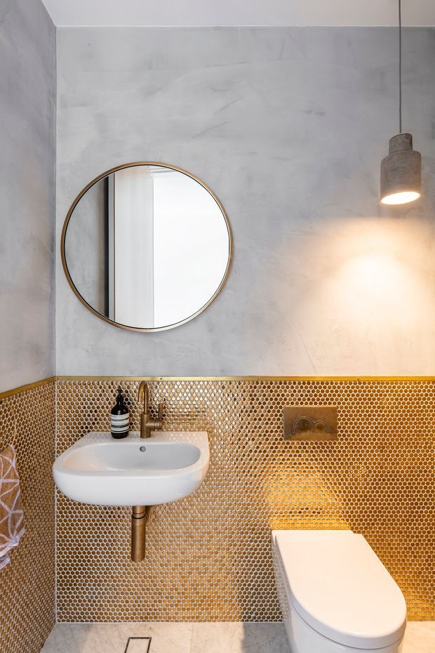 Compact contemporary bathroom featuring a wall-mounted white basin with exposed bronze-toned plumbing, a concealed-cistern toilet, round mirror with a thin brass frame, warm pendant light, pale textured plaster walls, and a striking golden-brown mosaic tile dado wrapping the lower walls. Light stone or concrete-look floor tiles complete the space.