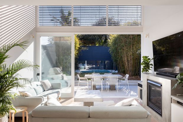 Bright contemporary living room with white walls, expansive floor-to-ceiling glazing and upper clerestory windows opening to a landscaped courtyard with a swimming pool and outdoor dining area. A large sectional sofa, fireplace, wall-mounted television, indoor plants, and light-toned furnishings create an airy indoor-outdoor setting.