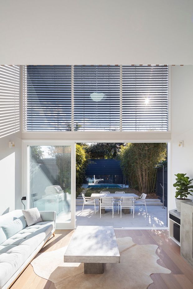 Bright, contemporary living room opening directly onto a paved outdoor dining terrace and garden courtyard. The room features expansive floor-to-ceiling glazing and upper horizontal Venetian blinds, white walls, pale timber flooring, a light upholstered sofa, sculptural pale coffee table, and abundant natural light. A dining setting and swimming pool are visible outside.