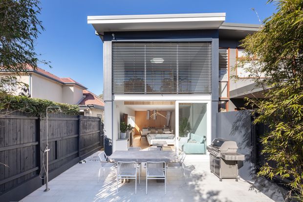 Exterior view of a contemporary multi-level residence with a flat roof, projecting upper-storey glazing fitted with horizontal blinds, and a glazed ground-floor living area opening onto a paved outdoor entertaining space. The terrace includes a dining setting and barbecue, with timber fencing and neighbouring homes visible around the compact urban site.