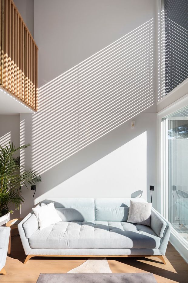 Bright double-height living room with a pale upholstered sofa, large floor-to-ceiling glazing, white walls, warm timber flooring, and a timber vertical-slat balustrade or screen at the upper level. Strong sunlight creates dramatic linear shadows across the walls and sofa, with indoor planting adding a natural touch.
