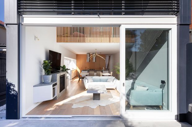 Bright contemporary living room viewed from the exterior through wide glazed doors. The compact open-plan space features pale timber flooring, white walls, a white sofa and low coffee table, a wall-mounted television with fixed media cabinetry, indoor plants, and a dining area beyond. Dark exterior framing and horizontal screening contrast with the light interior, while vertical timber battens form a feature balustrade or privacy screen above.