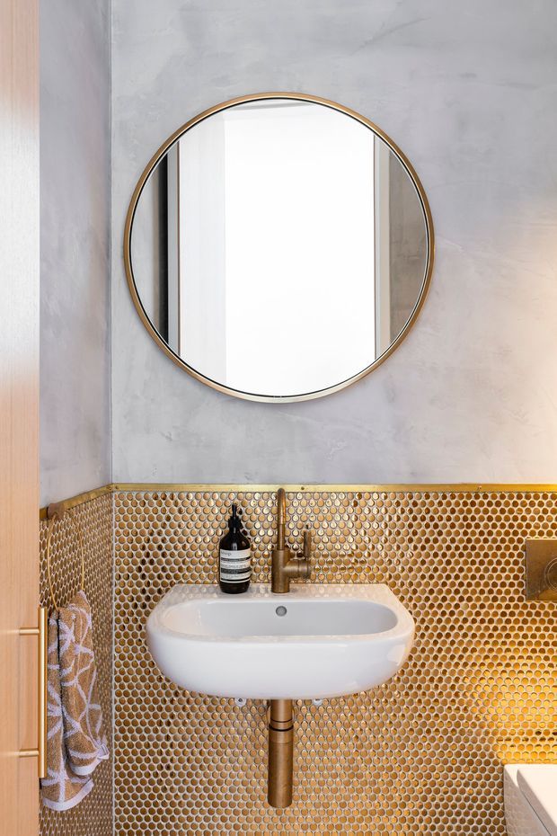 Compact contemporary bathroom featuring a wall-mounted white basin, exposed plumbing, a round mirror with a slim brass-toned frame, and a striking honeycomb mosaic tile wall in warm metallic gold and bronze tones. The upper wall has a pale grey textured finish, with timber cabinetry and a towel visible at the sides.