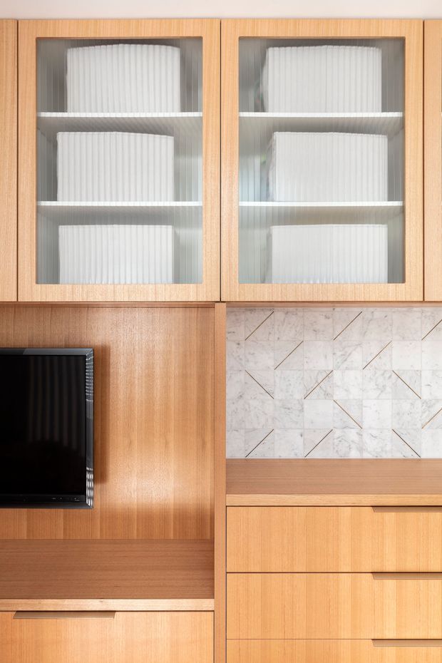 Close-up of a refined built-in timber entertainment and storage wall. Warm vertically grained wood cabinetry includes glass-front upper cupboards with white ribbed objects inside, a wall-mounted black television, open shelving, drawers, and a marble-look tiled feature backsplash with fine diagonal line details. The composition is highly geometric and focused on fixed joinery.
