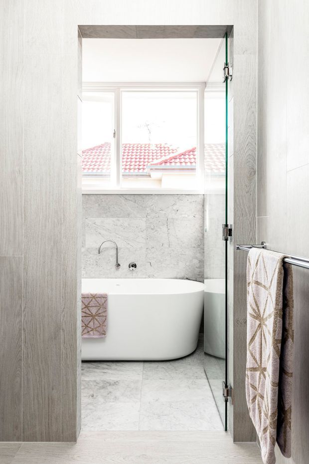 Bright contemporary bathroom viewed through a frameless glass door, featuring a freestanding white bathtub, pale marble-look wall and floor surfaces, a window, and a chrome towel rail with patterned towels. Vertical light wood-look wall panelling frames the opening, while a red tiled roof is visible through the window.