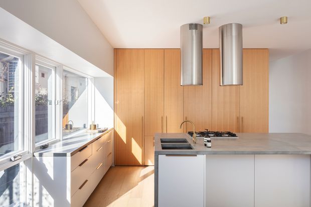 Bright contemporary kitchen with a large pale stone island, white flat-panel cabinetry, warm vertical timber wall cabinetry, and a bank of windows along the left side. Two cylindrical stainless-steel rangehoods hang above the island cooktop, while sunlight creates strong geometric shadows across the cabinetry and timber floor.