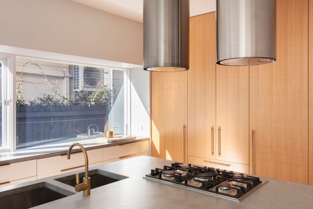 Bright contemporary kitchen featuring a large light-grey island with an inset dark double-bowl sink, brushed metal gooseneck tap, and built-in five-burner gas cooktop. Full-height warm timber cabinetry lines the rear wall, with two cylindrical stainless-steel pendant rangehoods above the island. A wide window provides natural light and views to a planted exterior courtyard or neighbouring property.