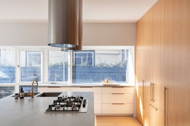 Bright contemporary kitchen with a large grey island and integrated gas cooktop, a cylindrical stainless-steel rangehood, white lower cabinetry, and full-height warm timber cabinetry along the right wall. Large windows provide abundant natural light and overlook neighbouring buildings.