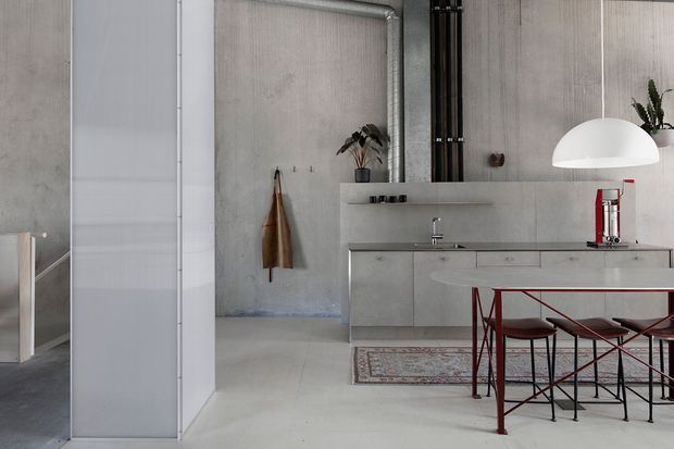 Open-plan minimalist kitchen with a pale concrete-look finish, handleless cabinetry, a long island with dark red metal bar stools, and exposed black vertical services or structural elements. A large white pendant light hangs over the island, while a hallway and adjacent spaces are visible to the left. The room has a restrained industrial character with neutral grey tones and sharp linear detailing.