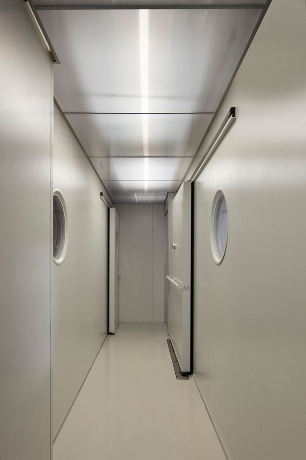 A narrow, highly sanitary white corridor with smooth panelled walls, a segmented reflective ceiling, circular porthole-style windows, sealed flooring, and linear drainage grates. The space appears utilitarian and controlled, resembling a cleanroom or industrial processing facility.