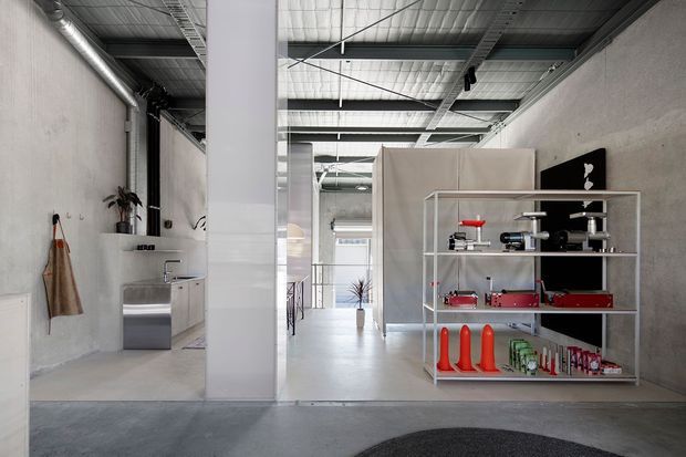 A minimalist industrial-style commercial interior with exposed metal ceiling structure, polished concrete flooring, raw concrete walls, and a large white structural column. The open space includes a compact kitchenette or utility area at left, a central glazed partition, and open metal shelving displaying products or equipment at right.