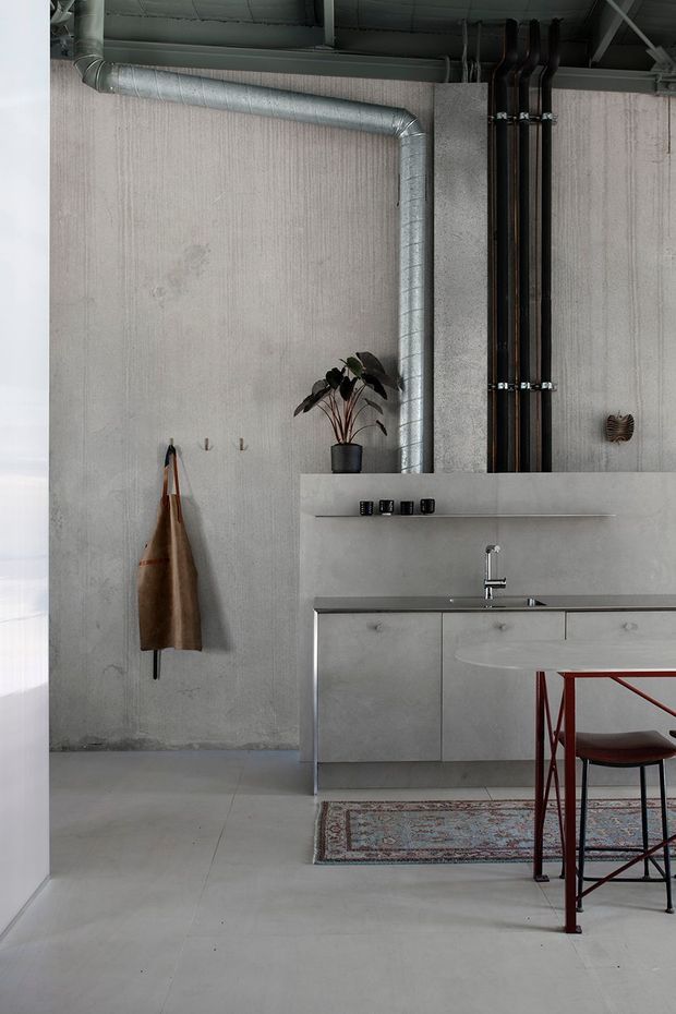 A compact industrial-style kitchen with exposed ductwork and dark plumbing pipes against a raw concrete wall and ceiling. The space features a minimalist grey concrete or cement-finished kitchen with integrated cabinetry, a slim benchtop, stainless tapware, a small plant, timber wall hooks, and a red metal-framed stool beside a patterned rug. The palette is predominantly cool grey with restrained red and brown accents.