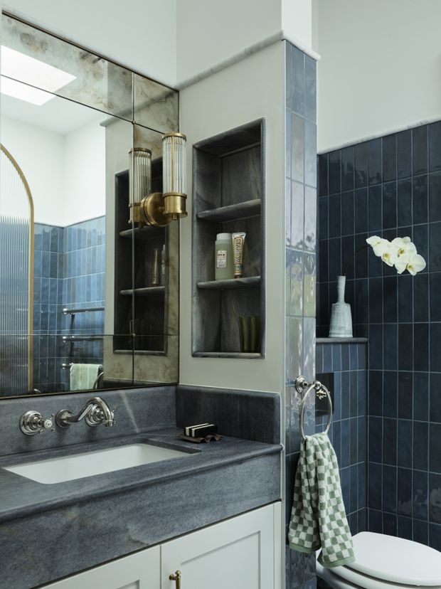 A refined bathroom interior featuring a dark blue tiled wall, a deep grey stone vanity benchtop with integrated white basin, pale cabinetry, mirrored wall panels, recessed open shelving, and polished brass wall-mounted tapware. A glass shower enclosure and bathtub are visible in the mirror reflection, with a small orchid and patterned hand towel adding decorative detail.