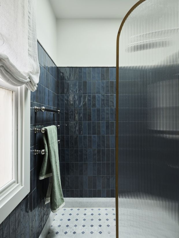 A compact, refined bathroom with deep blue tiled walls, a patterned blue-and-white floor, and a curved ribbed-glass shower screen framed in warm timber. A white-framed window with a fabric Roman blind provides natural light, while dark metal towel rails and a muted green towel add functional detail.