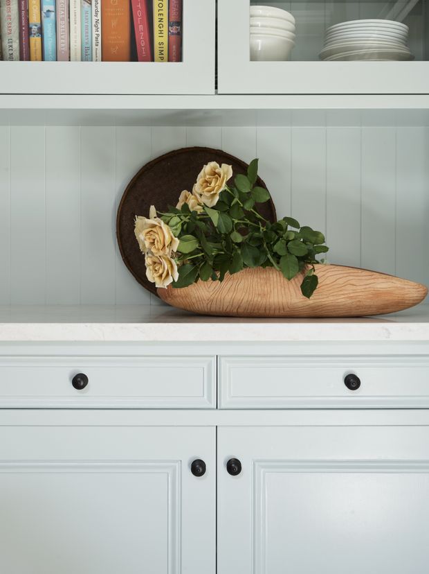 Close-up interior detail of a pale blue-grey built-in cabinet with raised-panel doors and drawers, dark round knobs, a white stone countertop, and open shelving above. The shelves display books, white crockery, and a decorative wooden bowl filled with flowers against vertical painted wall panelling.