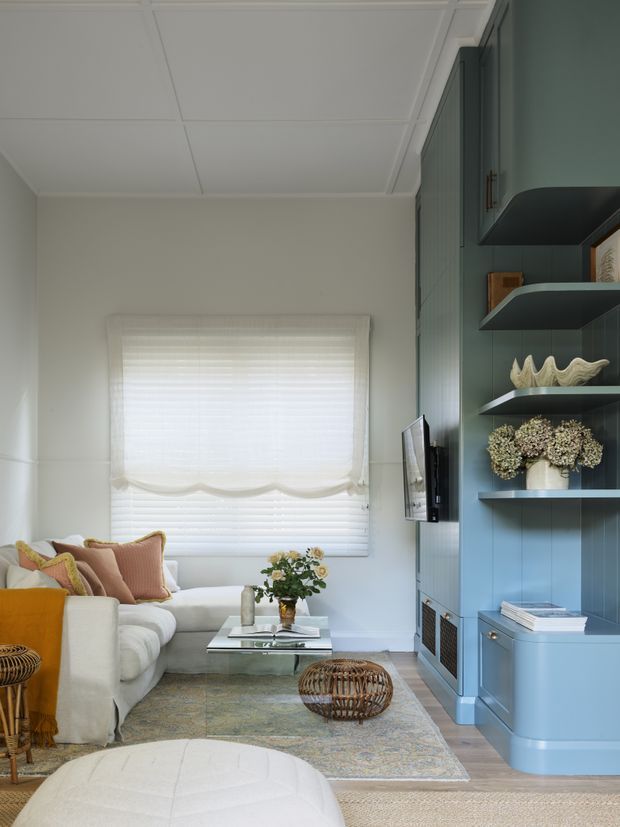 A compact, refined living room with a cream sectional sofa, layered neutral and blush cushions, a glass coffee table, patterned area rug, woven ottoman, and a pale upholstered foreground ottoman. A large window has a white Roman blind, while the right wall features built-in blue-green cabinetry, open display shelving, a wall-mounted television, and decorative objects. The room has a coffered-style panelled ceiling and a light, softly layered palette.