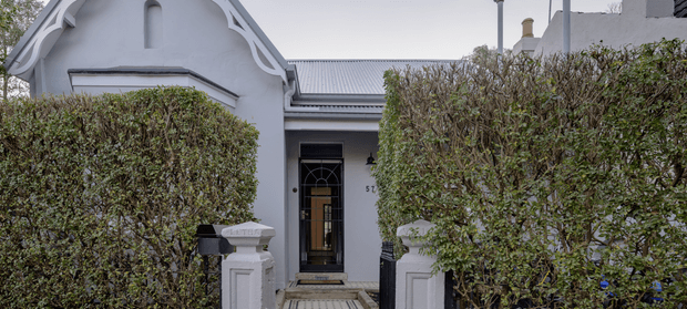 Front exterior view of a compact heritage-style residence, partly screened by carefully trimmed hedges. The pale rendered facade features an ornate gabled entrance with decorative timber detailing, a dark glazed front door, masonry entry pillars, and a corrugated metal roof.