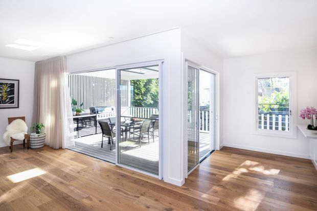 Bright, open residential living area with warm timber flooring, white walls and ceiling, large sliding glass doors, and expansive glazing opening onto a furnished outdoor terrace or balcony. Minimal furnishings include a lounge chair, side table, plant, artwork, and a small table with chairs visible outside. The space feels airy and contemporary, with strong indoor-outdoor connection and abundant natural light.