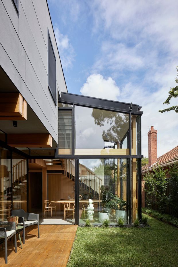 Exterior view of a contemporary multi-level residential extension with dark-framed floor-to-ceiling glazing, a glazed link or atrium, grey panel cladding, timber-lined soffits, and a timber deck opening onto a small landscaped lawn. A warm timber interior with a visible stair is seen through the glazing, while an adjacent brick chimney and tiled roof appear at the right.