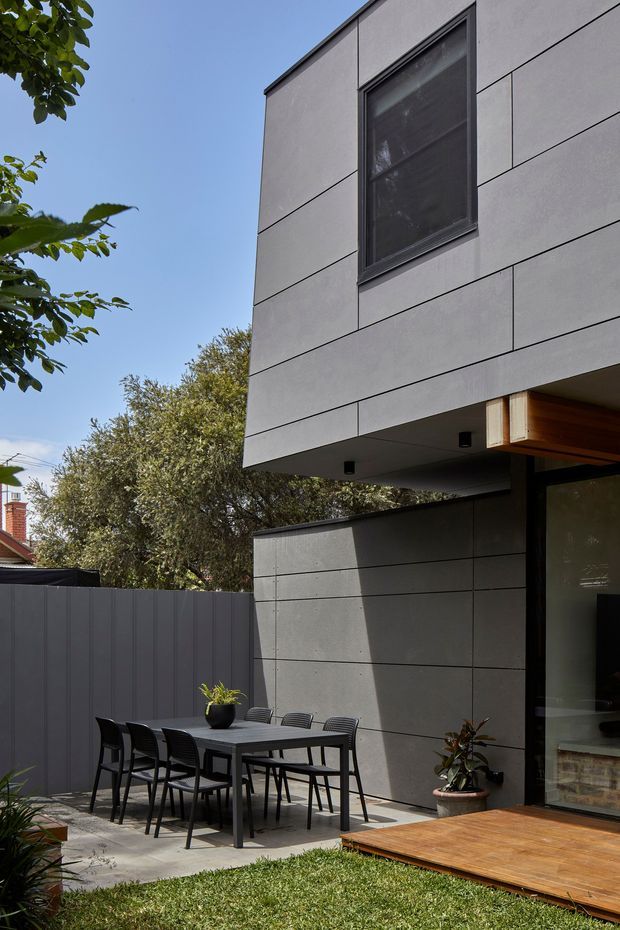 A modern two-storey residential exterior with a projecting upper volume clad in large grey fibre-cement panels, dark-framed window, and a partially enclosed grey-panelled courtyard. The ground-level outdoor dining area includes a dark rectangular table and chairs, with a timber deck and glazed opening beneath a warm timber soffit. Established trees and a privacy fence surround the compact garden.