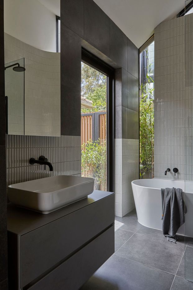 A refined contemporary bathroom featuring a freestanding white bathtub, a wall-mounted black tap and shower set, and a white vessel basin on dark built-in vanity cabinetry. Vertically ribbed light tiles cover the walls, contrasted by large dark wall panels and large-format grey floor tiles. Full-height glazing and a narrow outdoor opening bring in natural light and greenery.