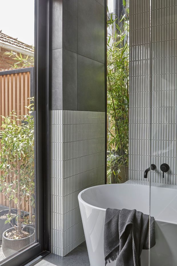 A contemporary bathroom featuring a white freestanding bathtub beside a large black-framed window. The walls are clad in narrow vertical ribbed grey tiles, paired with dark charcoal wall panels and matte black wall-mounted tapware. A planted courtyard and timber screening are visible outside, bringing greenery and natural light into the space.