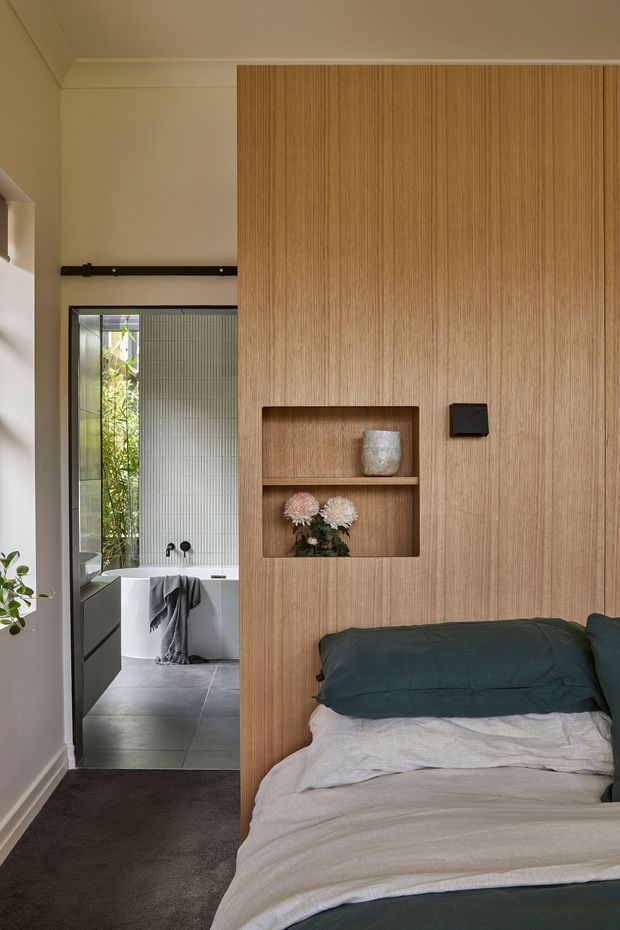 A compact, warm-toned bedroom viewed toward an adjoining bathroom. The bedroom features a floor-to-ceiling vertical timber-panelled wall with integrated open display niches, a partially visible bed with dark teal and light bedding, dark carpet, and simple white ceiling cornices. A black-framed opening with a sliding barn-style rail leads directly to a bathroom containing a freestanding white bathtub, grey tiled floor, and textured glazed wall treatment. The overall design is restrained, contemporary, and softly natural.