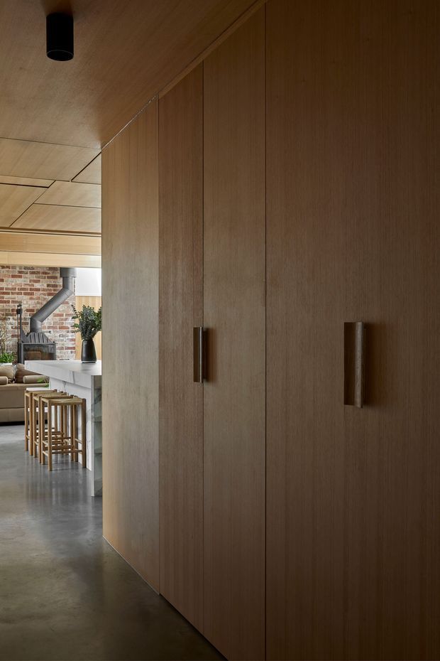 Interior view of a warm, contemporary open-plan kitchen and living area, dominated by a full-height wall of dark timber veneer cabinetry with long vertical handles. The space includes a concrete floor, timber-lined ceiling, white kitchen island with stools, exposed brickwork, and industrial-style ducting. A black cylindrical ceiling light adds contrast.