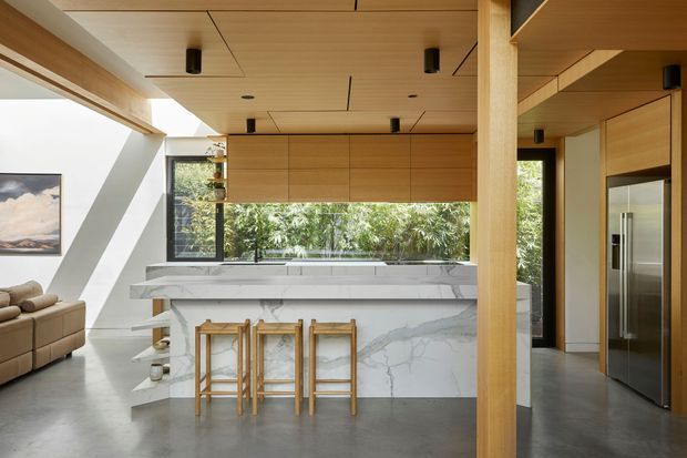 A bright, open-plan contemporary kitchen with a large marble-look island and waterfall sides, three timber bar stools, extensive light timber cabinetry, and a matching timber ceiling. Full-height glazing brings in greenery, while a living area with a sofa is visible at left. A prominent timber structural post separates the kitchen from a corridor or adjoining space.