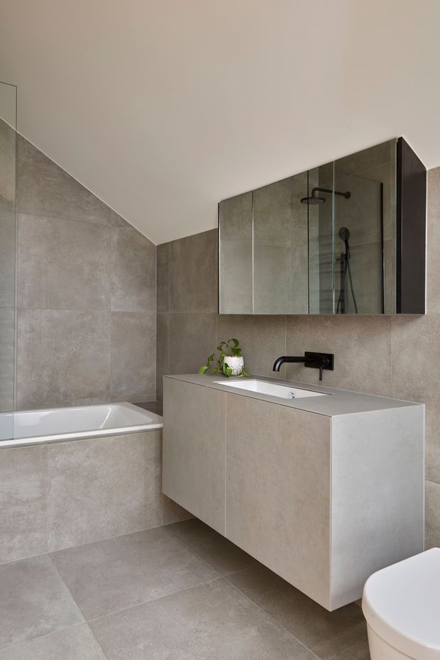 A contemporary bathroom with a wall-mounted floating vanity, integrated white basin, matte black wall-mounted tap, large frameless mirrored cabinet, and a glass-enclosed shower visible in the reflection. Large grey-beige stone-look tiles cover the floor and walls. A white bathtub sits beneath a sloping ceiling, with a partial toilet visible at the lower right. The room has a restrained, minimalist material palette and clean geometric lines.