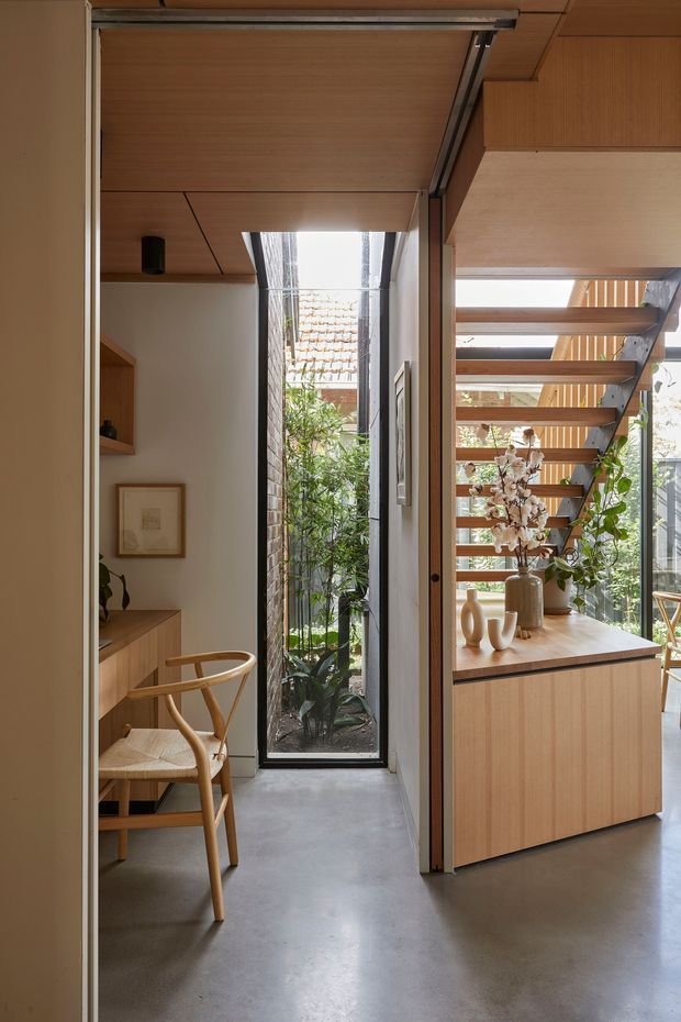 A warm, timber-lined interior hallway or transitional space with a compact built-in home office nook on the left and a timber cabinet beneath an open-riser staircase on the right. A tall narrow glazed opening brings in daylight and views of an internal planted courtyard. The space features polished grey concrete flooring, extensive light timber cabinetry and ceiling lining, black-framed glazing, and a sculptural stair with timber treads and dark metal supports.