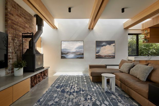 A warm contemporary living room with exposed timber ceiling beams, a full-height exposed brick fireplace wall, and a black freestanding wood-burning stove. A large brown sectional sofa faces the fireplace and sits over a patterned blue-grey rug, with a small white side table in front. Built-in timber cabinetry, recessed black spotlights, framed landscape artwork, and large windows overlooking greenery complete the space.