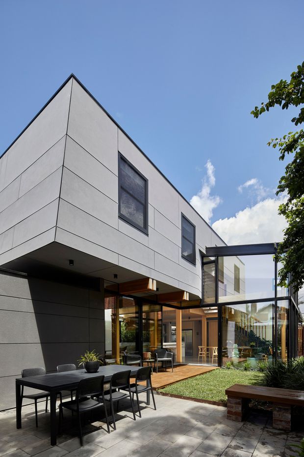 Exterior view of a contemporary multi-level residence arranged around a landscaped courtyard. The home features crisp light-coloured panel cladding, strong geometric forms, dark-framed glazing, a projecting upper volume, and a glazed connection or passage. A paved courtyard includes an outdoor dining setting, timber decking, lawn, planting, and a built-in bench.