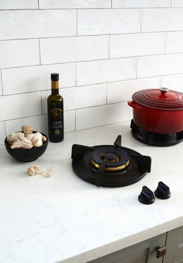 Close-up kitchen scene featuring a black built-in gas cooktop set into a pale stone benchtop, with black control knobs, a red lidded Dutch oven, white subway-tile splashback, garlic, and an olive oil bottle. The image focuses on the cooking surface and surrounding kitchen finishes.