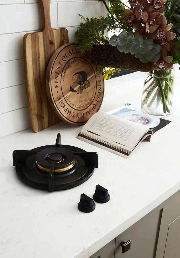 A styled kitchen vignette featuring a black gas cooktop with brass detailing and separate black control knobs set into a white engineered-stone benchtop. White subway-tile splashback, grey cabinetry with dark hardware, timber cutting boards, a round wooden serving board, an open cookbook, and a vase of flowers create a warm domestic setting.