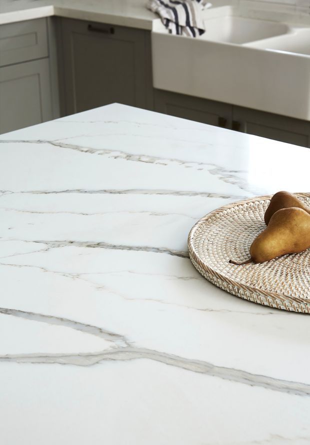 Close-up of a bright kitchen featuring a large white marble-look island benchtop with prominent grey veining. Grey cabinetry and a deep white farmhouse-style sink are visible in the background, with a woven tray holding pears on the island.