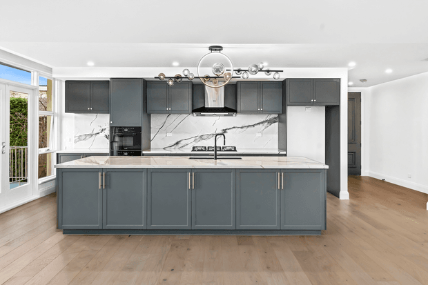 Bright contemporary kitchen featuring a large charcoal-grey island with shaker-style cabinetry, a pale marble-look stone benchtop and matching full-height splashback. The rear wall includes integrated appliances, gas cooktop, rangehood and dark cabinetry. Light timber flooring, white walls, large glazed doors, and a statement globe pendant create an airy, refined interior.
