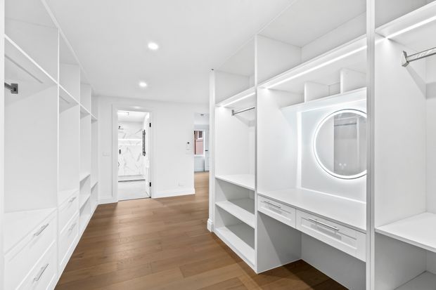 Bright, spacious walk-in wardrobe with extensive white built-in cabinetry, open shelving, drawers, hanging rails, and an illuminated round mirror integrated into a dressing vanity. Warm timber flooring contrasts with the crisp white joinery, and a bathroom is visible through the doorway in the background.